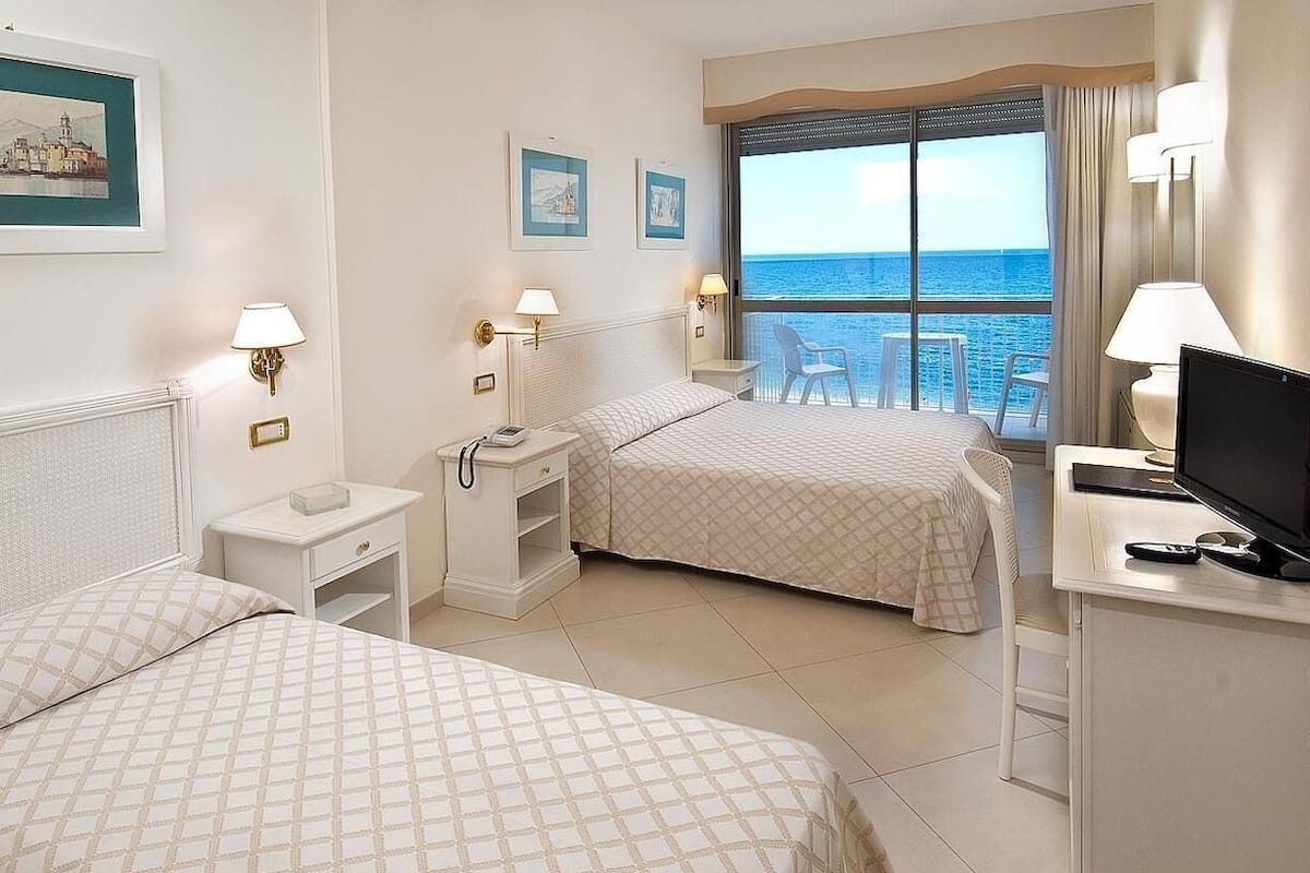 A hotel room with two beds, a desk with a TV, and a balcony with a view of the sea.