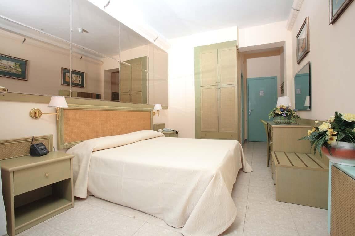 Quarto de hotel com cama, mesas de cabeceira, espelho, escrivaninha com luminária e arranjo de flores.