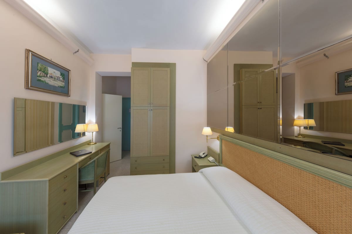 A hotel room with a bed, a desk with a lamp, a telephone, a mirror, and a wardrobe.