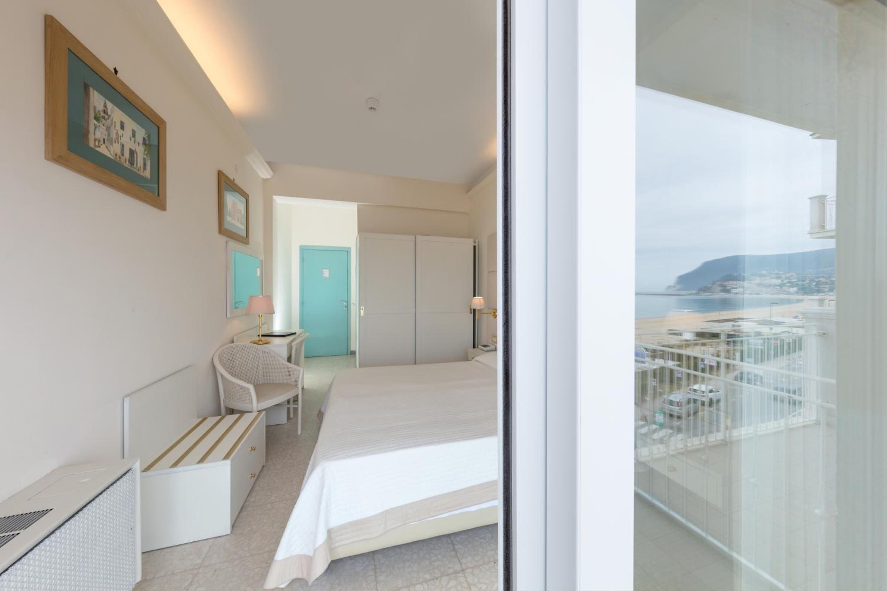 Quarto de hotel com cama, cadeira, escrivaninha e vista para a paisagem urbana através de uma varanda.