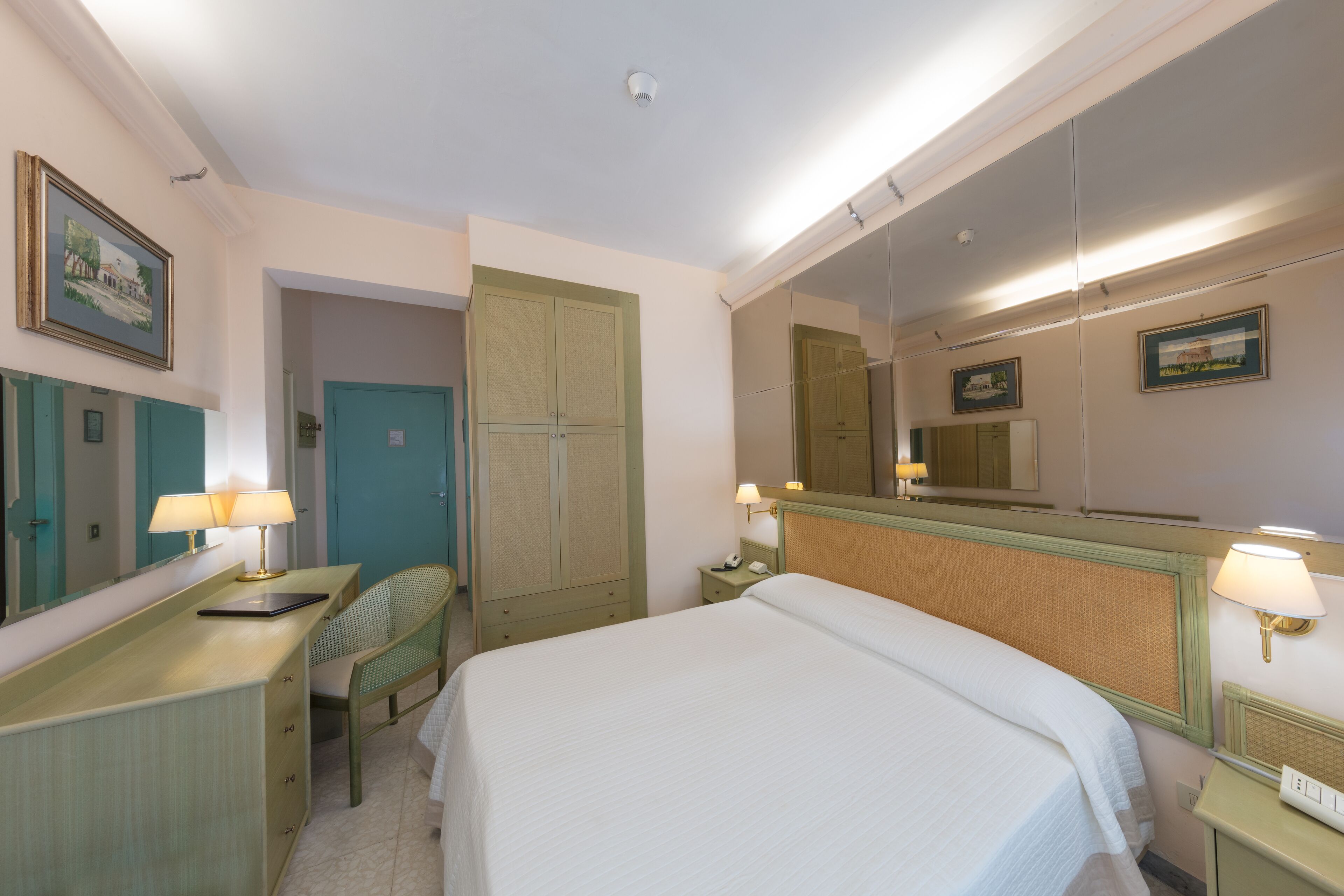 Quarto de hotel com cama, escrivaninha com luminária, cadeira, armário e espelho.