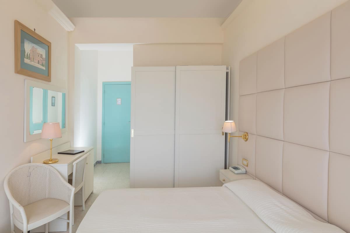 A bedroom with a bed, a desk with a lamp, a chair, a wardrobe, and a nightstand with a phone.