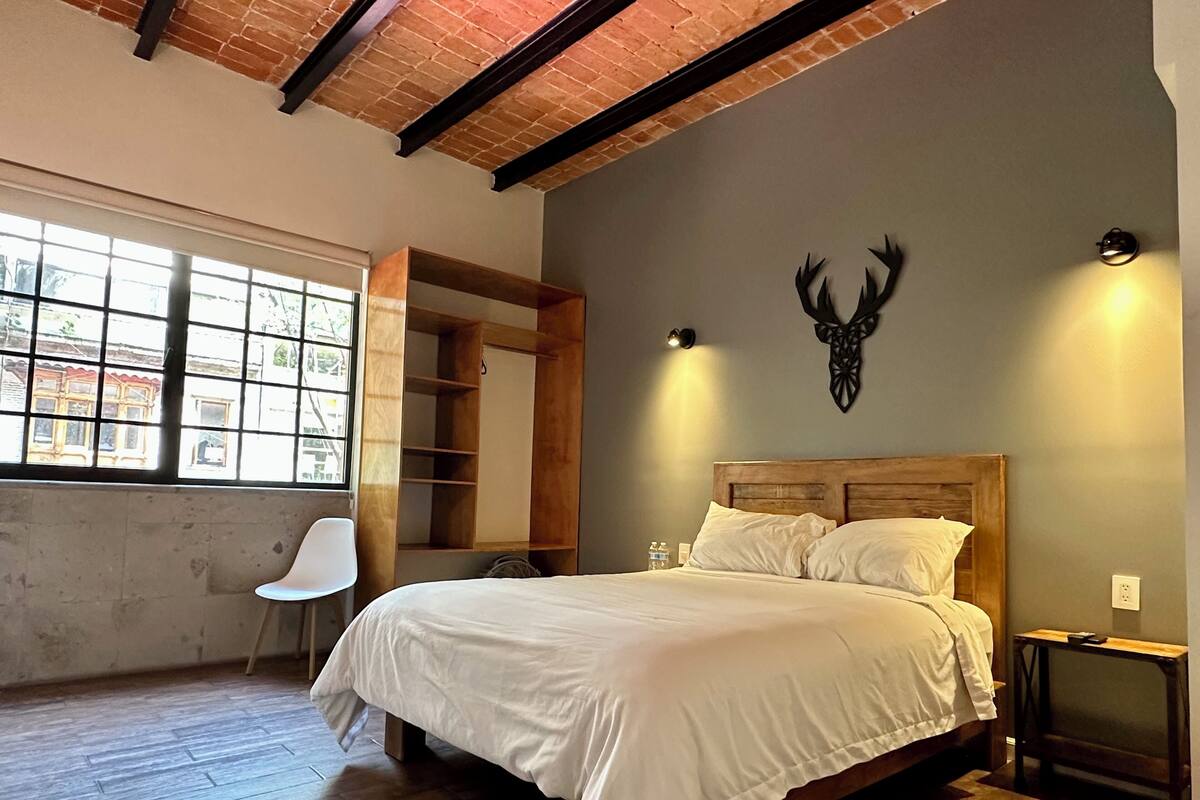 A bedroom with a large bed, a wooden headboard, a wall-mounted deer head decoration, a window with a view of buildings, and a built-in wooden wardrobe.