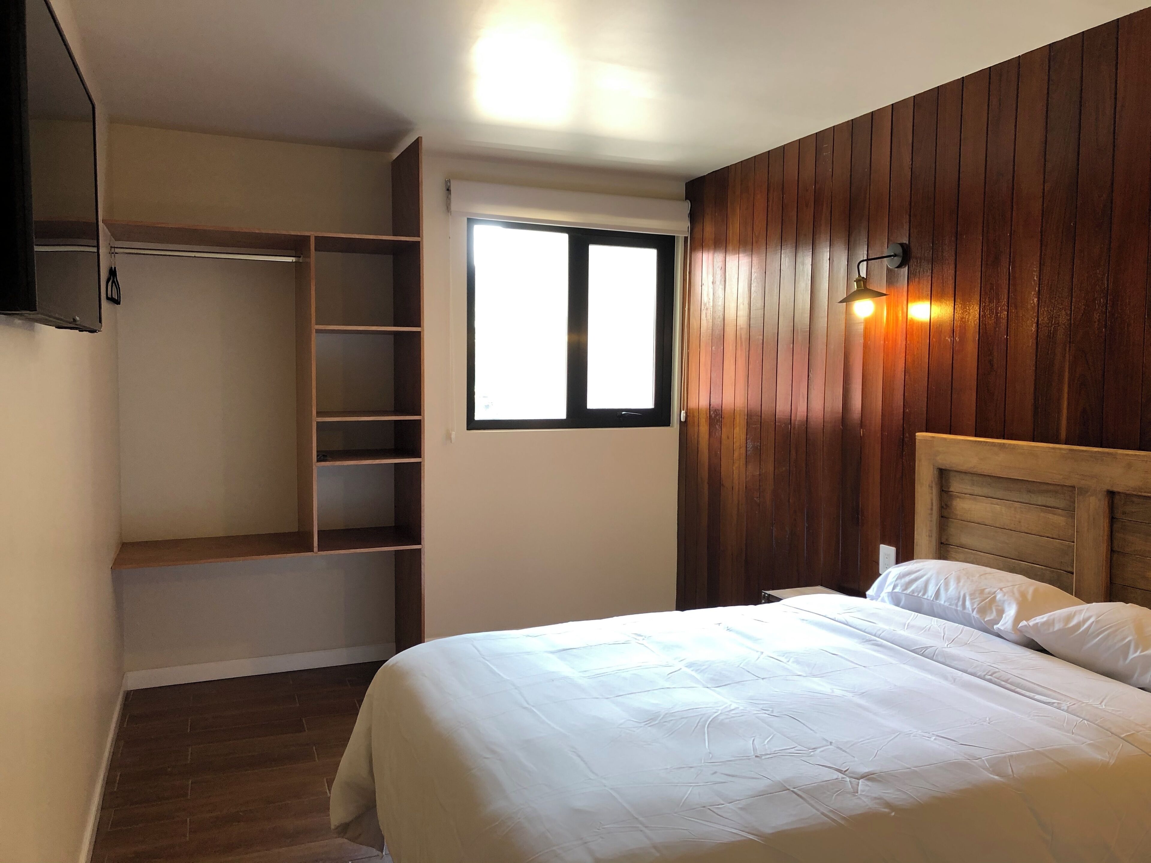 A bedroom with a bed, a wooden headboard, a wardrobe with shelves, a window, and a wall-mounted light fixture.