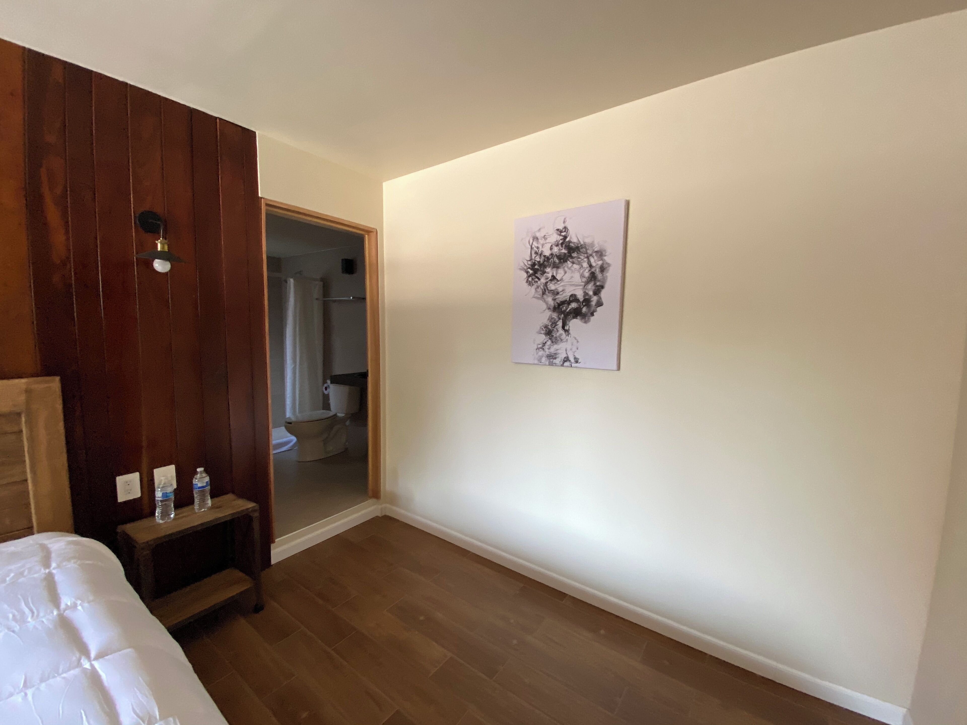 A bedroom with a wooden headboard, a bed, a nightstand with water bottles, and a bathroom visible through an open door.