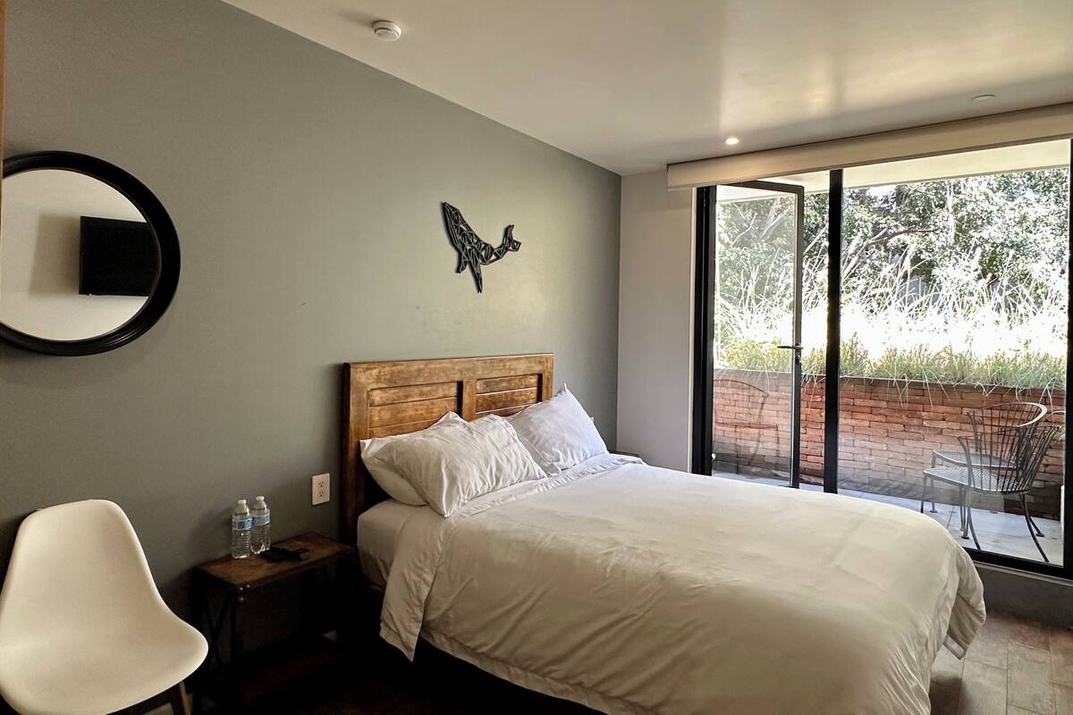 A bedroom with a large bed, a bedside table with a lamp, a chair, and a view of the outdoors through a sliding glass door.