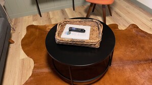 Een ronde tafel met een gevlochten mand waarin een afstandsbediening en een boekje liggen, op een kuitenvachttapijt.
