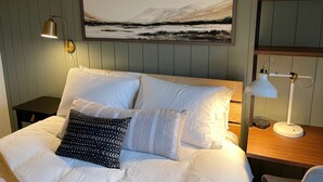 Een net opgemaakt bed met witte lakens en gestreepte kussens, een aan de muur hangend landschapsschilderij, een bedlamp en een houten nachtkastje.