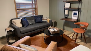 Een woonkamer met een bank, twee fauteuils, een salontafel, een bijzettafeltje, een boekenkast en een raam met jaloezieën.