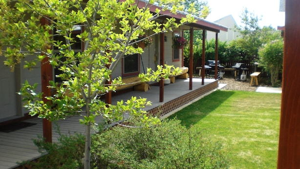 Een overdekt terras met houten leuning, een kleine tuin met gazon en een gebouw op de achtergrond.