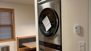 Een wasserette met twee gestapelde voorladers, wasmachines en drogers, een bankje, een houten bankje, een raam met zonwering en een kleine prullenbak.