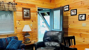 A cozy cabin interior with wooden walls, a leather chair, a blue sofa, a dining table, and framed fish pictures on the wall.