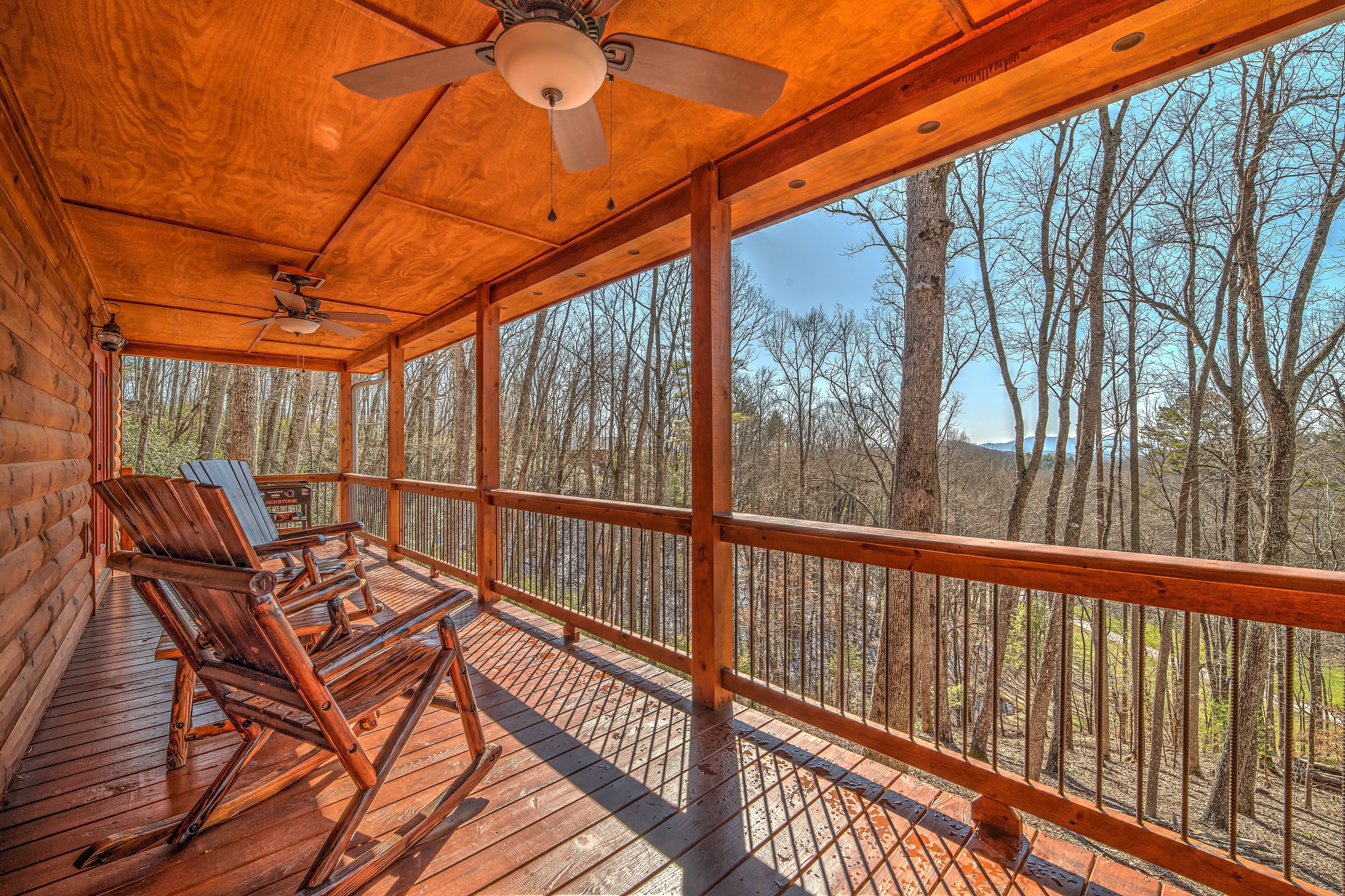 A wooden deck with rocking chairs and a ceiling fan, overlooking a forested area.