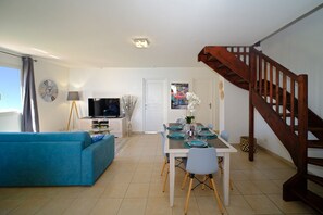 Un salon avec un canapé bleu, un coin repas avec une table dressée pour quatre personnes, un téléviseur posé sur un meuble et un escalier en bois.