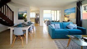 Un salon moderne avec un canapé bleu, une table à manger et des chaises. Un escalier se trouve à gauche, et de grandes fenêtres offrent une vue sur l’océan.
