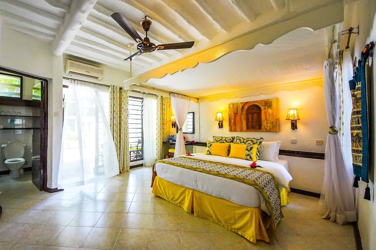 Habitación de hotel con cama, ventilador de techo, baño y vistas al exterior.