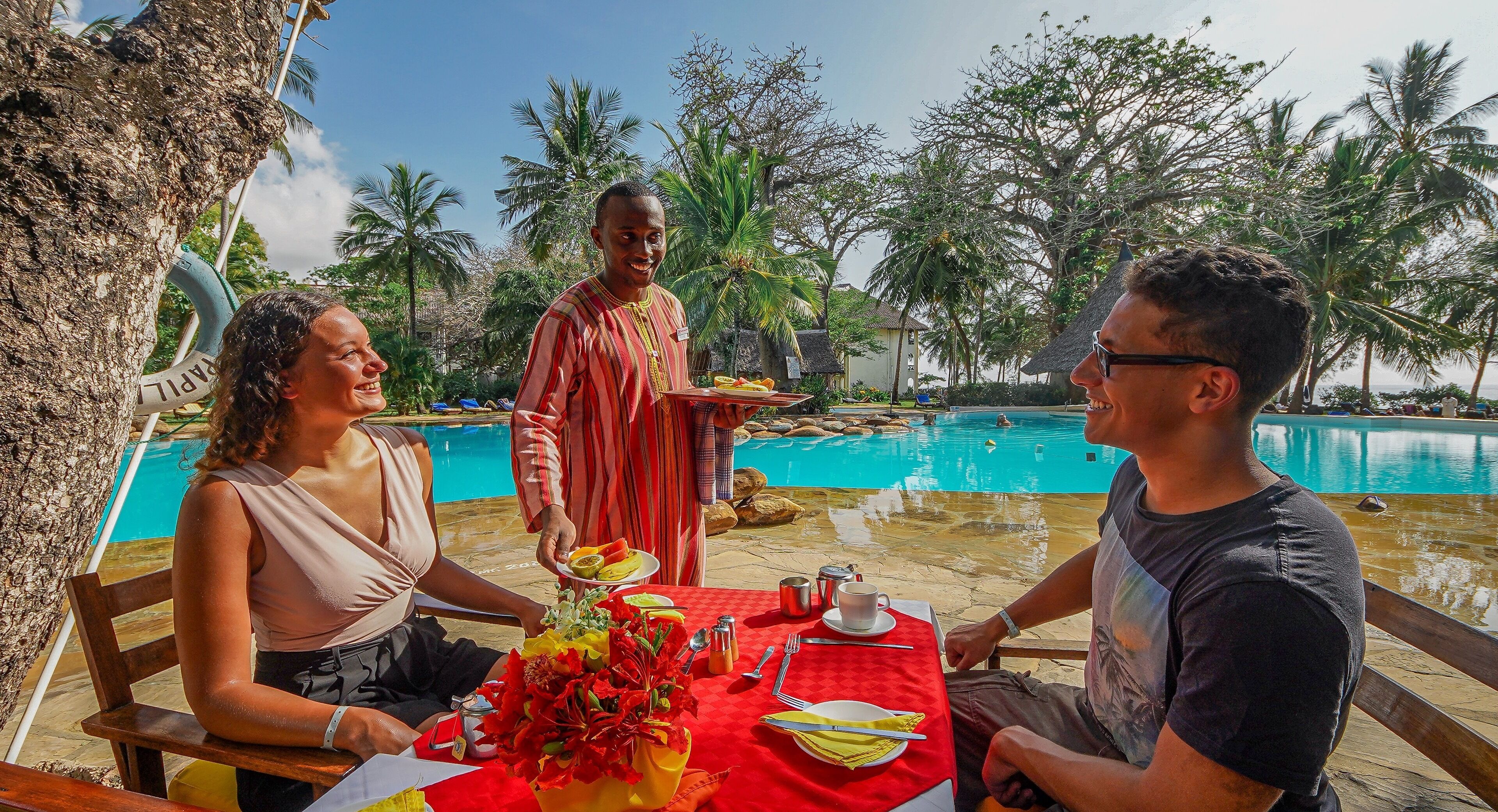 A dining table with a red tablecloth, a vase of red flowers, and a pool in the background.