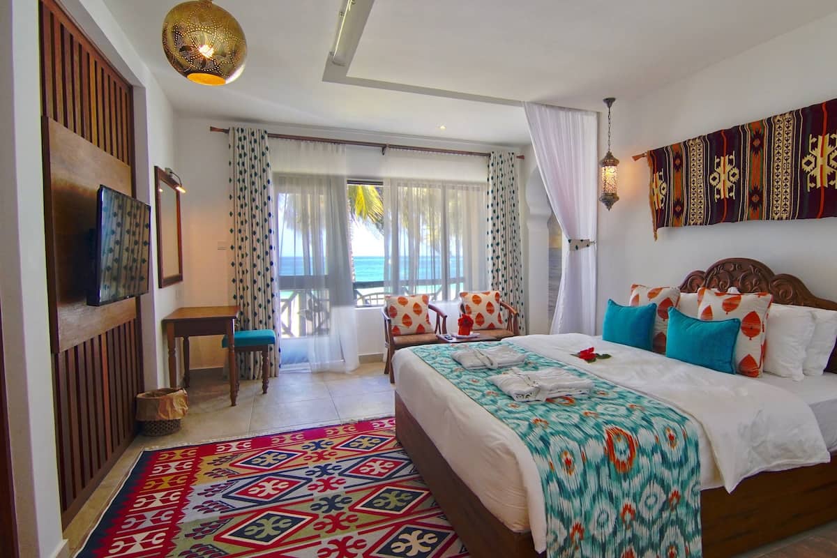 Una habitación de hotel con una cama grande, televisión, balcón con vista al mar y alfombra estampada.