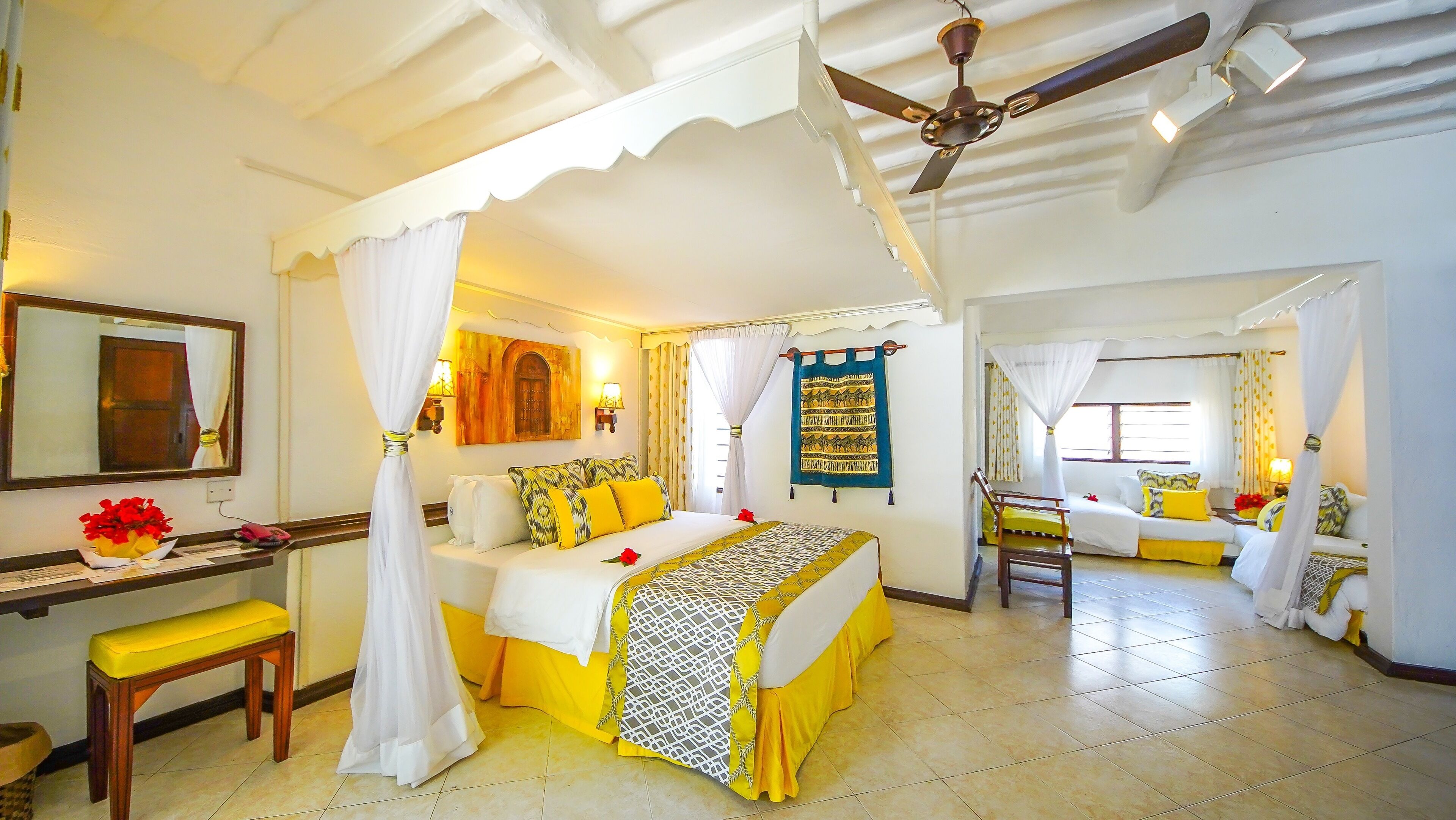 A room with a bed, a bench, a desk, a ceiling fan, and a window with curtains.