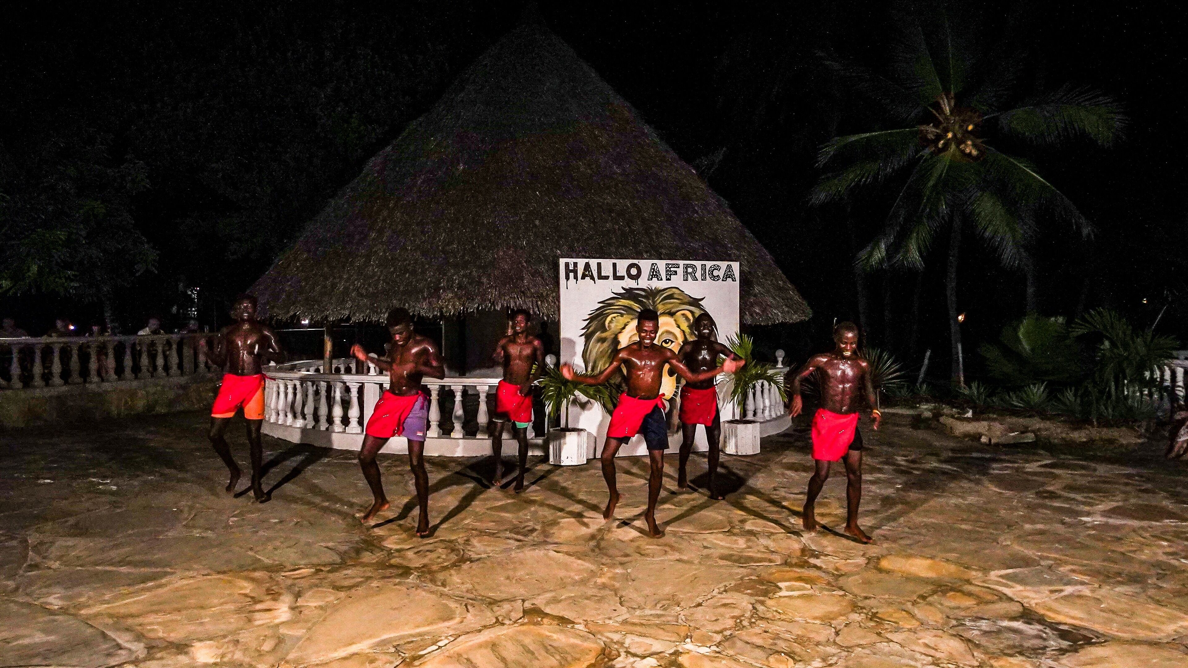 A group of people performing in front of a thatched-roof structure with a sign reading "HALLO AFRICA".