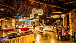 A restaurant with a high ceiling, stone pillars, and hanging lanterns. Tables are set with red tablecloths and various dishes. - Papillon Lagoon Reef (Diani Beach)