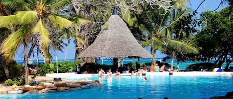A pool area with a thatched-roof structure surrounded by palm trees and a view of the ocean.