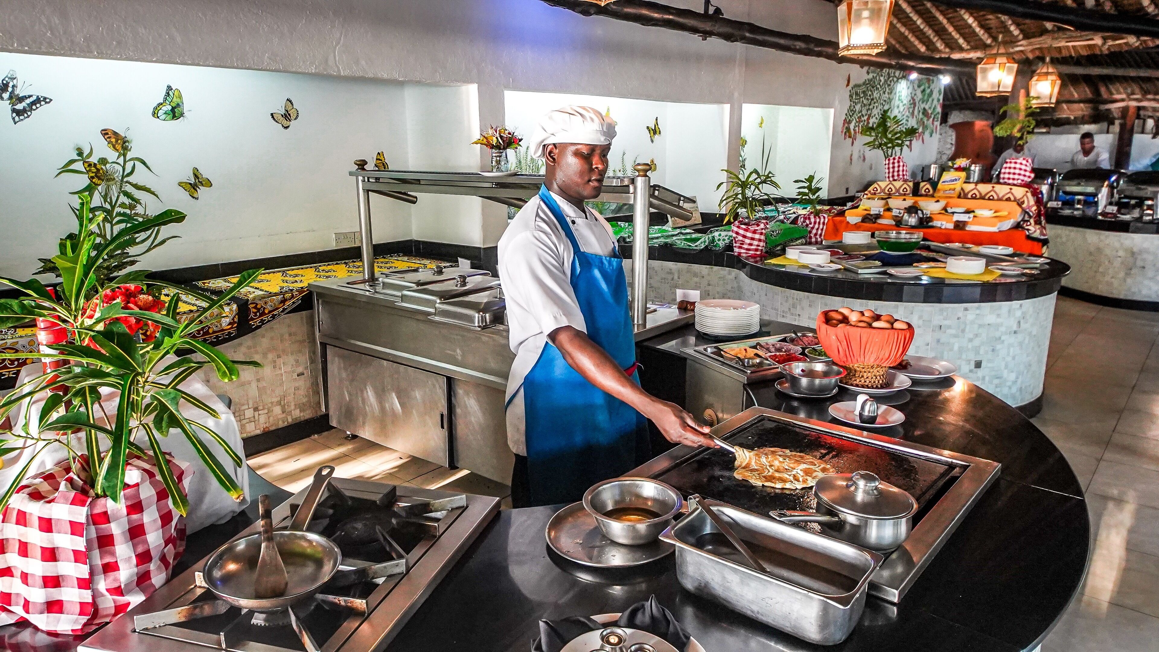 A chef in a kitchen with a buffet setup, including a variety of food items and a decorative plant.