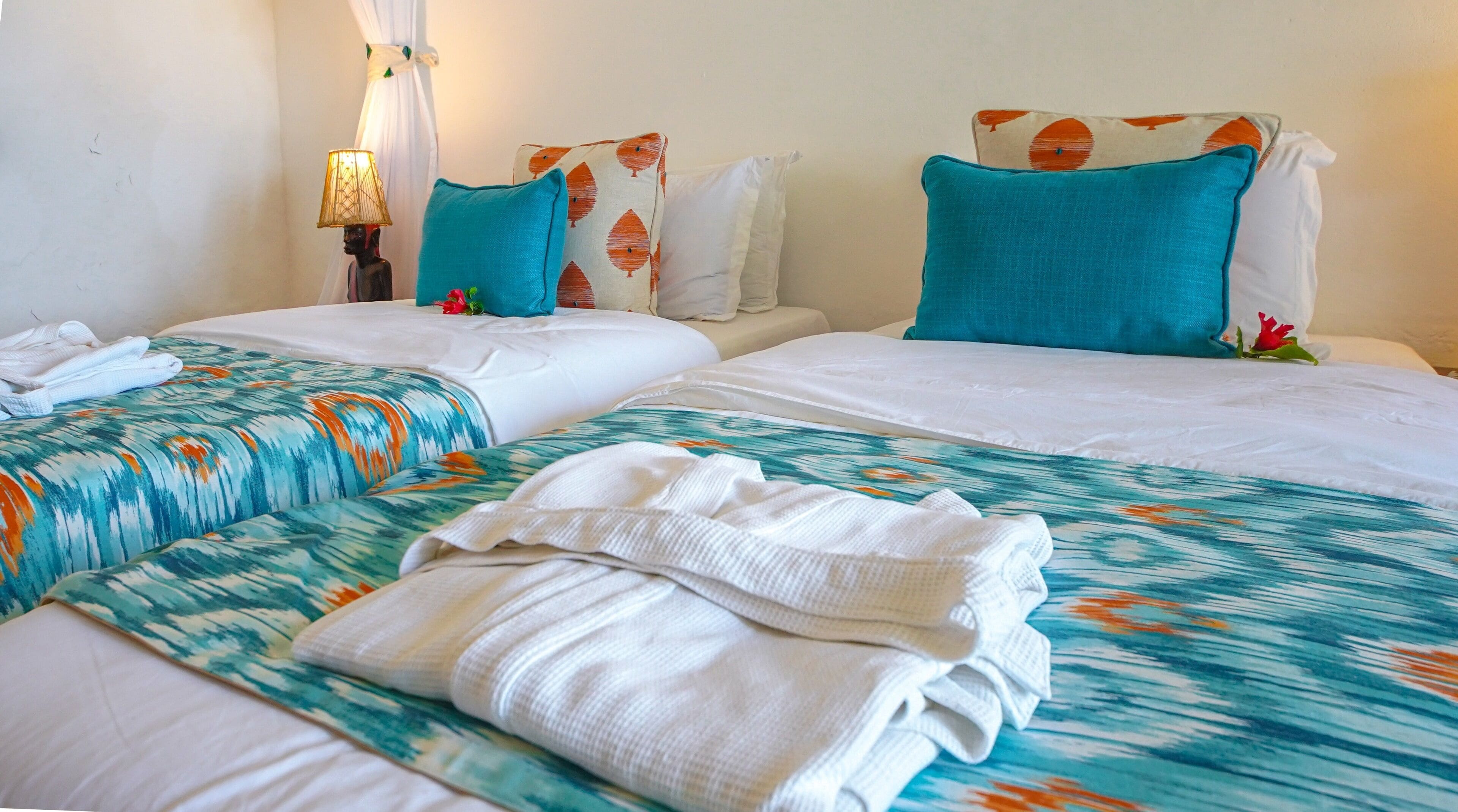 Une chambre d’hôtel avec deux lits, chacun recouvert d’une couvre-lit à motifs turquoise et orange, agrémentée d’oreillers blancs et d’une tête de lit blanche. On y trouve une table de chevet avec une lampe et un objet décoratif.