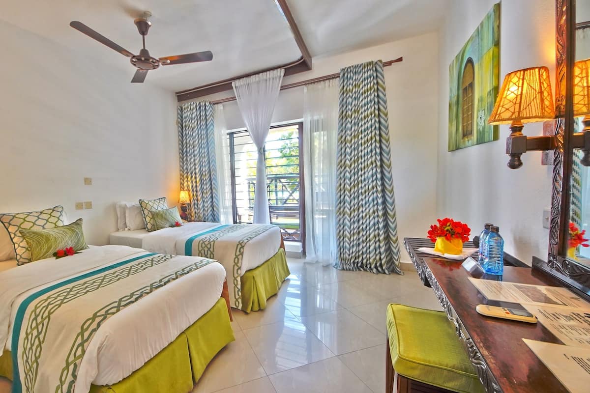 Habitación de hotel con dos camas, un escritorio con lámpara, ventilador de techo y vistas al exterior.