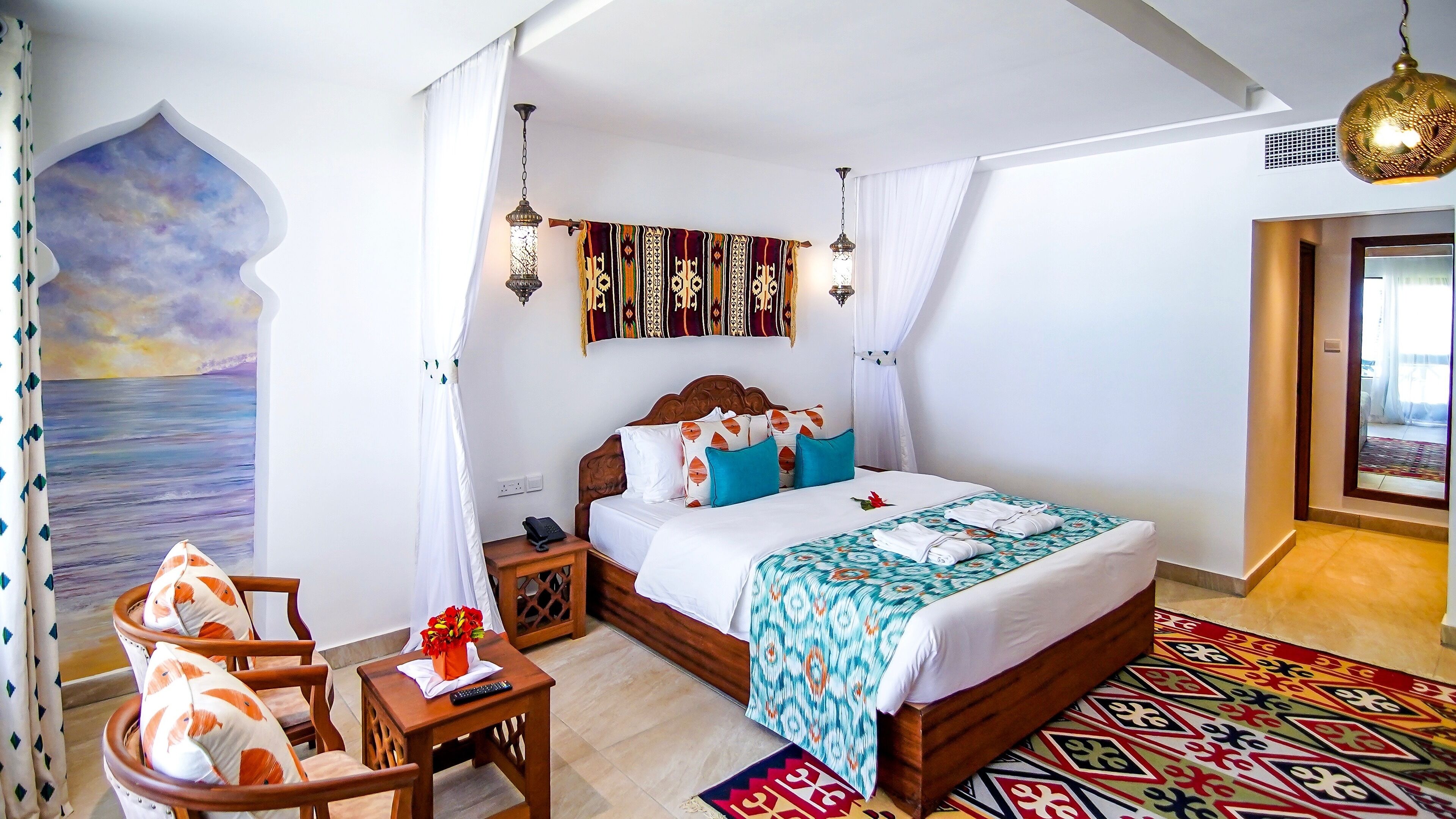 A well-lit bedroom with a bed, bedside tables, a nightstand, a chair, a patterned rug, and a wall mural.