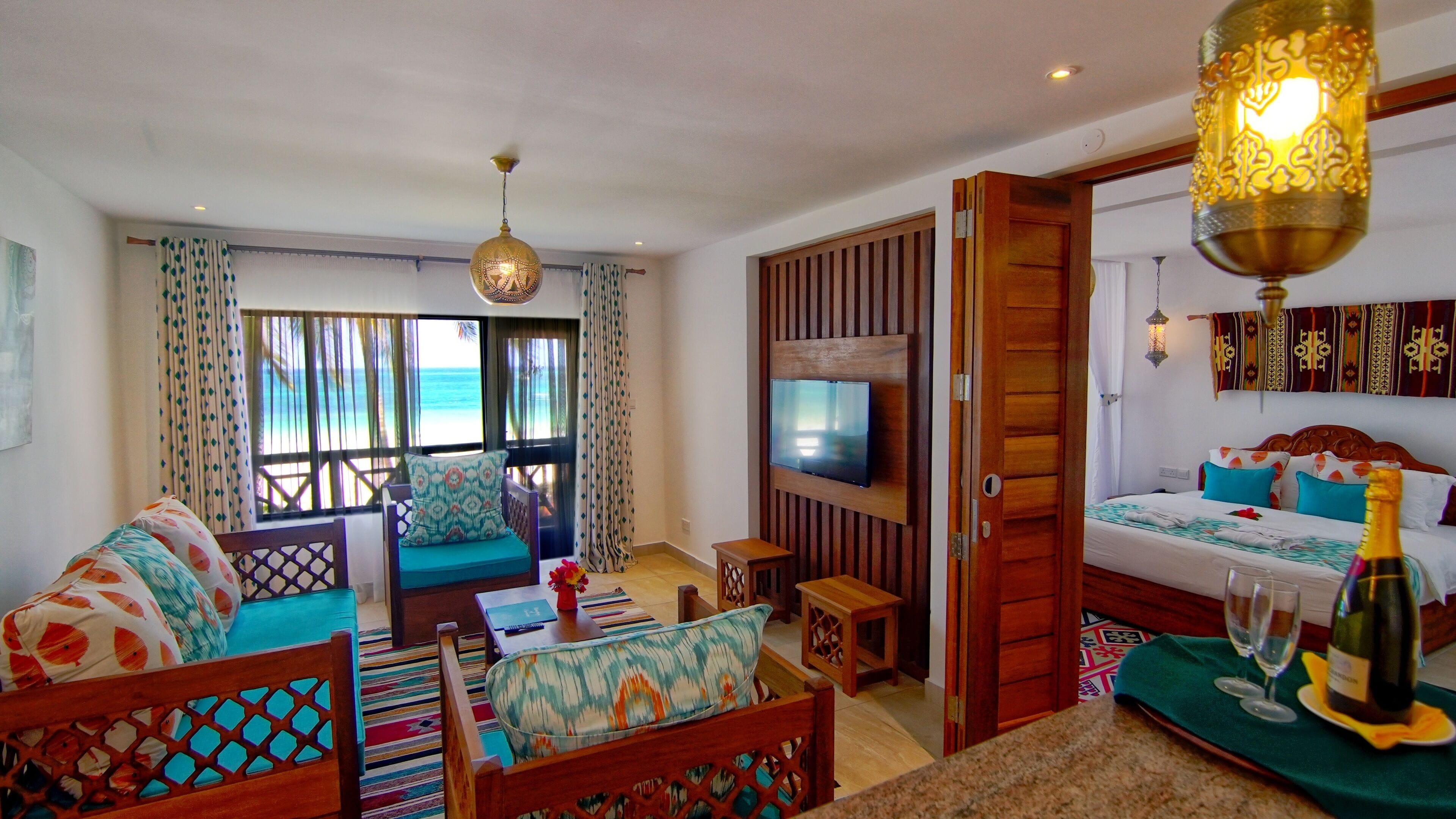 A hotel room with a sofa, a TV, a bed with colorful pillows, a wooden door, and a view of the beach.