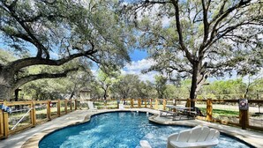 A swimming pool with a wooden deck, surrounded by trees and a fenced area. - Cozy and secluded Log Cabin near the Frio River with a private pool! (Concan)