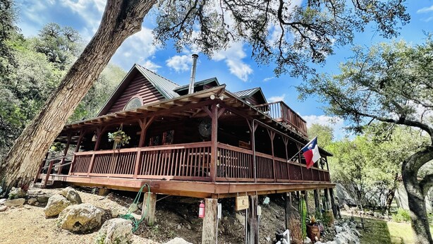 A rustic cabin with a wooden deck, a chimney, and a flag. - Cozy and secluded Log Cabin near the Frio River with a private pool! (Concan)