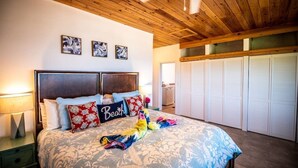 Ein Schlafzimmer mit Holzboden, einem Bett mit floralem Überwurf und einem Kissen mit der Aufschrift 'Beach'. An der Wand hängen drei gerahmte Bilder und es gibt einen Schrank mit weißen Türen.
