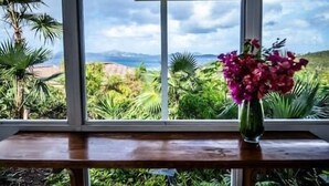 Un comedor con una mesa y dos sillas, un florero con flores, y vistas a vegetación exuberante y montañas distantes a través de amplios ventanales.