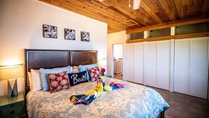 A bedroom with a wooden ceiling, a bed with a floral bedspread, a 'Beach' pillow, and a closet with white shutters.