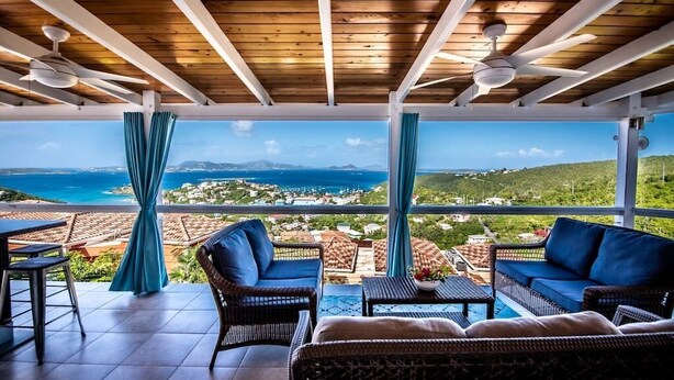 A covered outdoor seating area with wicker furniture, a pool table, and a view of the sea and landscape.