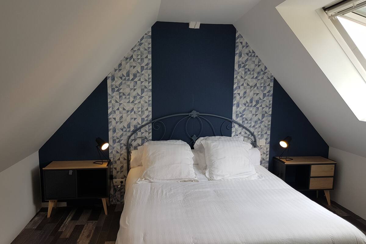 A bedroom with a sloped ceiling, a bed with white bedding, two bedside tables with lamps, and patterned wallpaper.