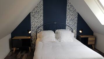Quarto com teto inclinado, cama com roupa de cama branca, duas mesas de cabeceira com abajures e papel de parede estampado.
