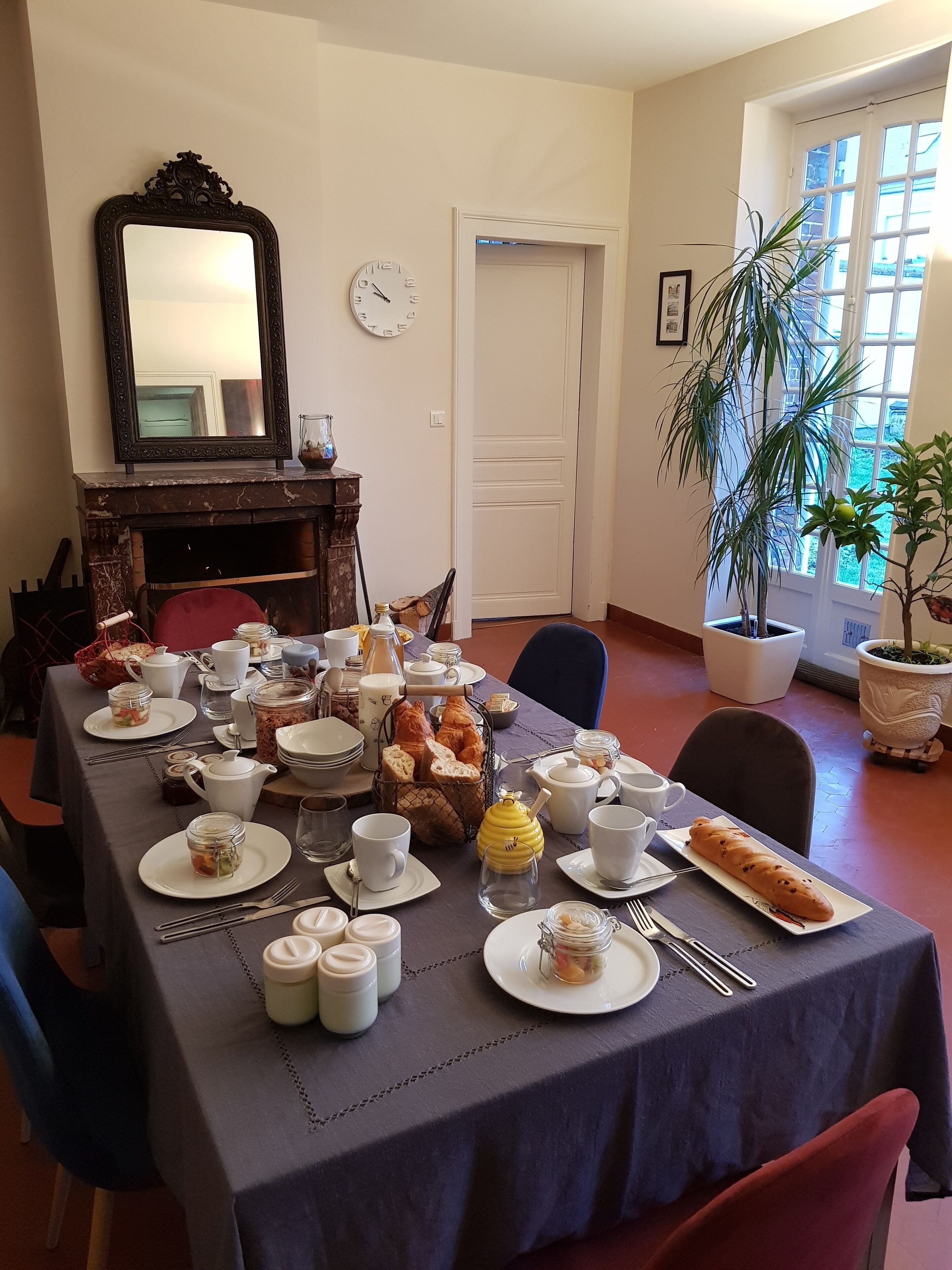 Un coin repas avec une table mise pour le petit-déjeuner, comprenant du pain, des viennoiseries et du thé. On y trouve également une cheminée, un miroir et une fenêtre agrémentée de plantes.