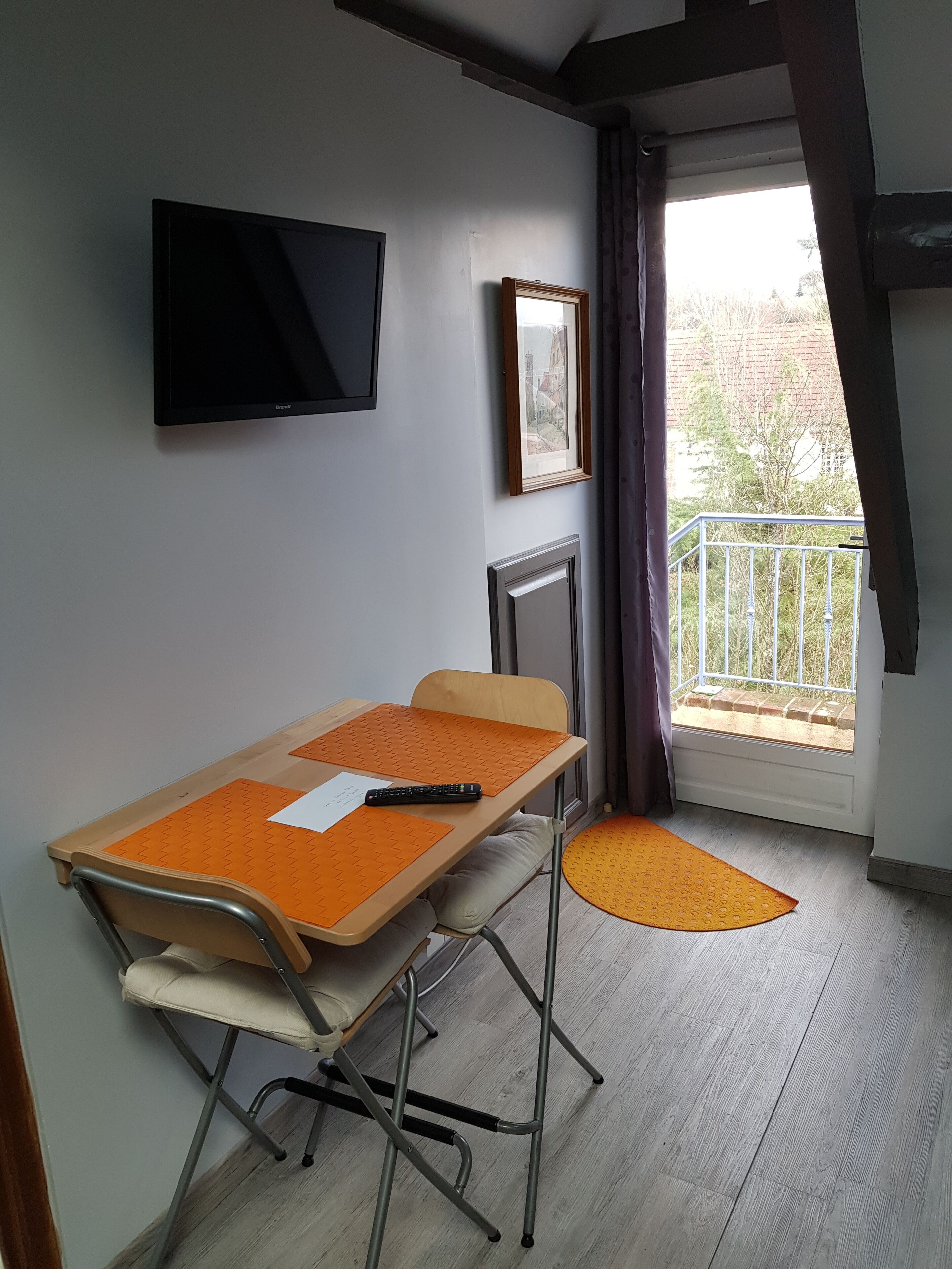 Een kamer met een uitschuifbaar bureau, een stoel, een aan de muur bevestigde televisie, een lijst met een afbeelding en een balkon met uitzicht op gebouwen en bomen.
