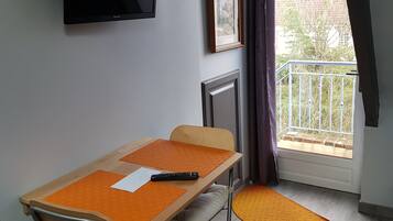 Een kamer met een uitschuifbaar bureau, een stoel, een aan de muur bevestigde televisie, een lijst met een afbeelding en een balkon met uitzicht op gebouwen en bomen.