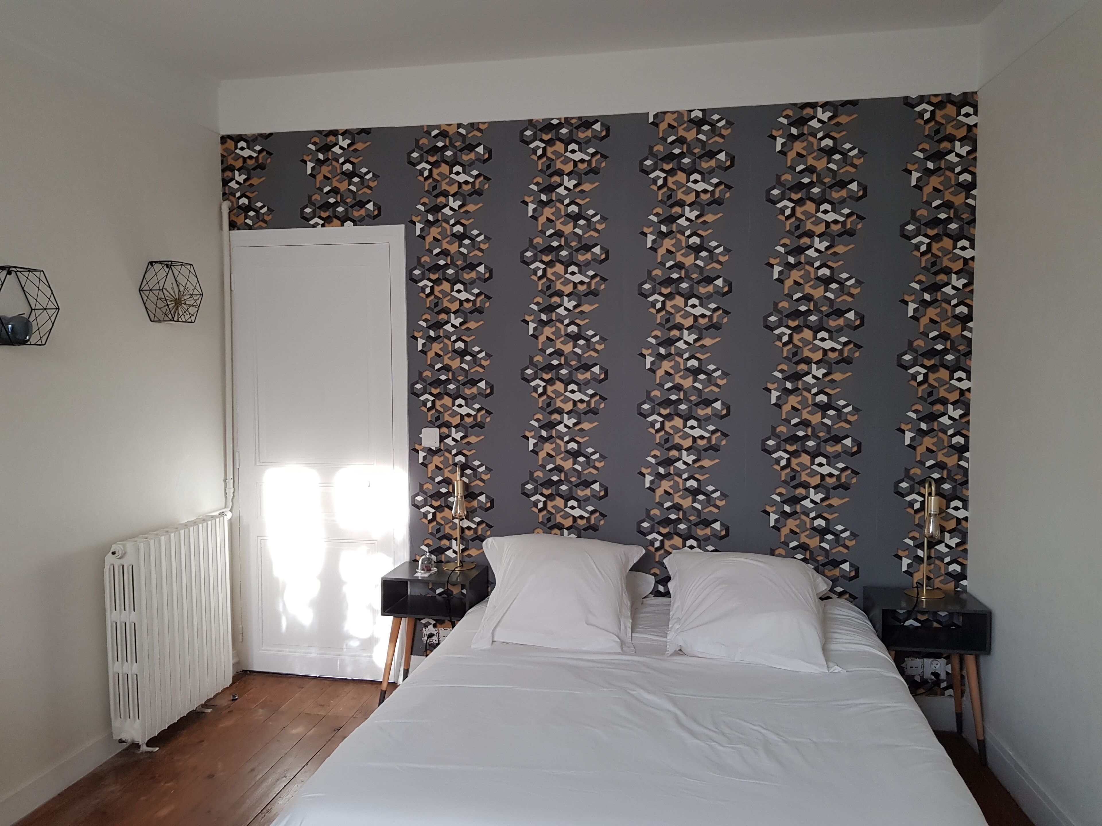 Un dormitorio con papel tapiz de diseño geométrico, una cama blanca, dos mesitas de noche y un radiador.