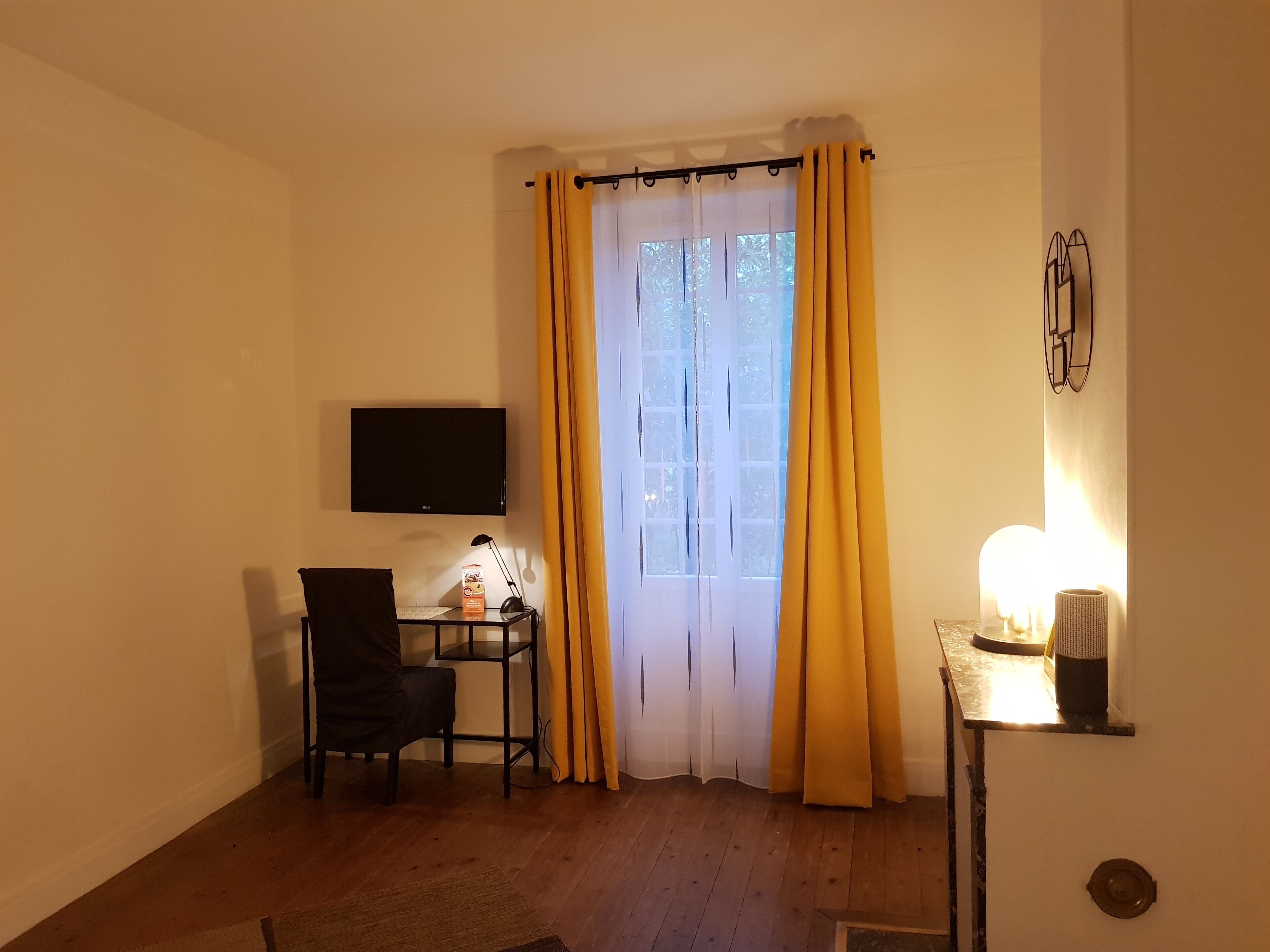 Una stanza con televisione a schermo piatto, una scrivania con lampada, una sedia e una finestra con tende gialle.