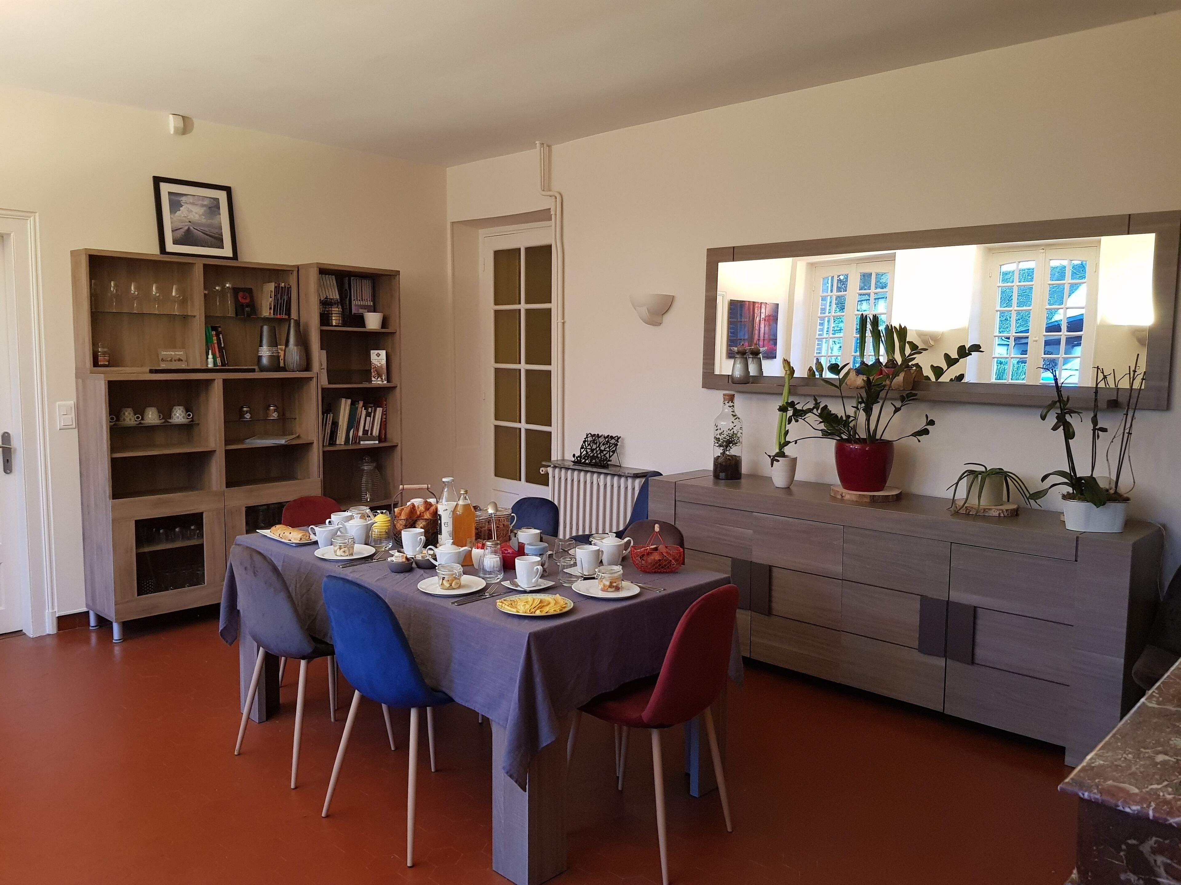 Un comedor con una mesa preparada para cuatro personas, rodeada de sillas. Hay una estantería a la izquierda y un aparador con objetos decorativos a la derecha.