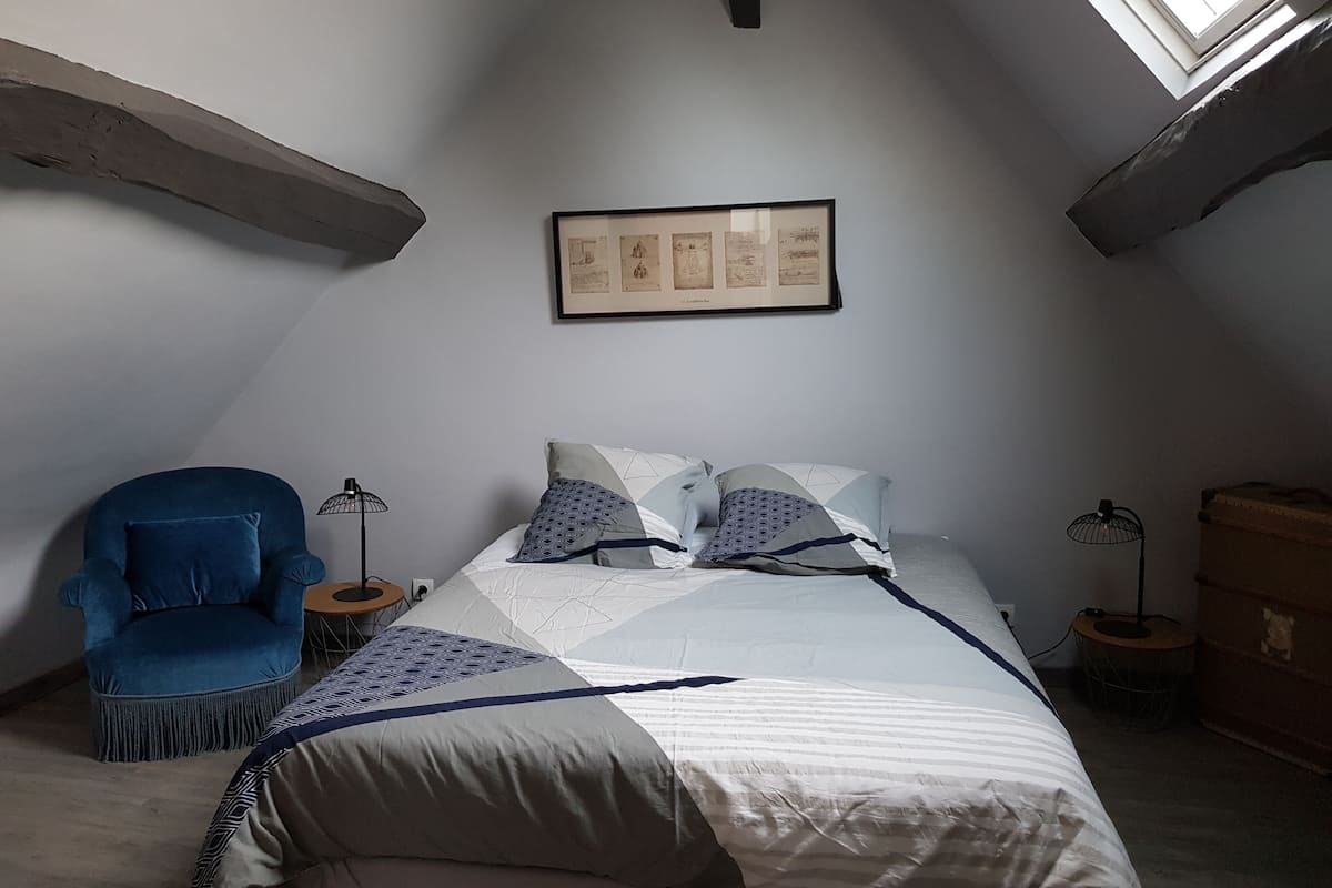 A bedroom with a sloped ceiling, a framed picture on the wall, a bed with patterned bedding, a blue armchair, and two floor lamps.