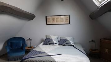 A bedroom with a sloped ceiling, a framed picture on the wall, a bed with patterned bedding, a blue armchair, and two floor lamps.