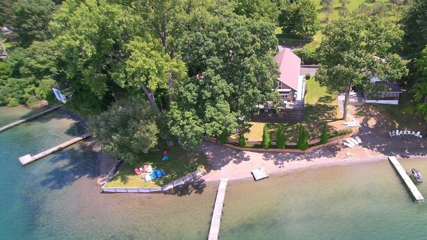 Vista aérea de una propiedad junto al lago con un muelle, un área verde con mobiliario exterior y una casa con techo rojo.