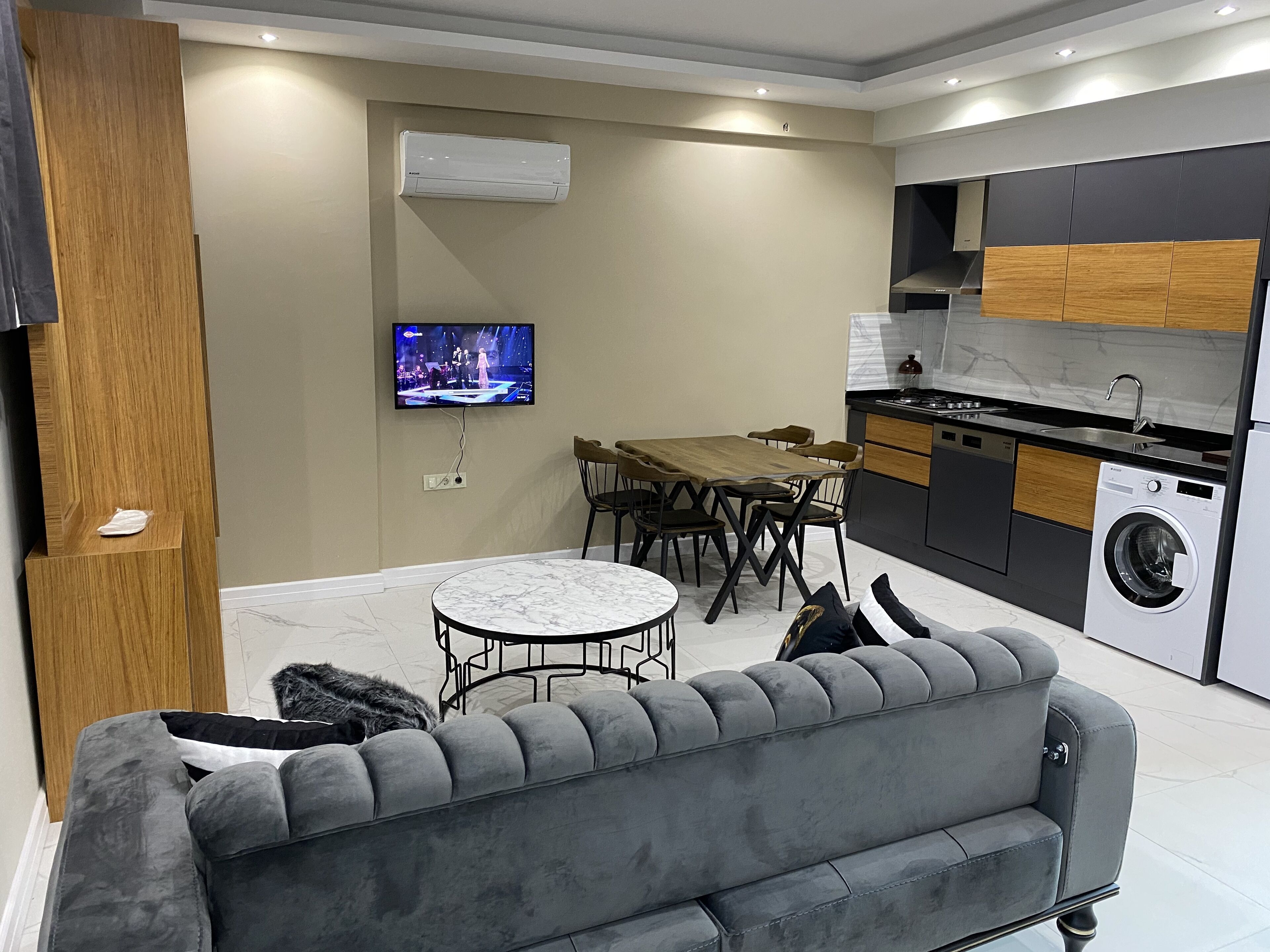 A modern kitchen with a built-in washing machine, a dining area with a marble table, and a grey sofa.