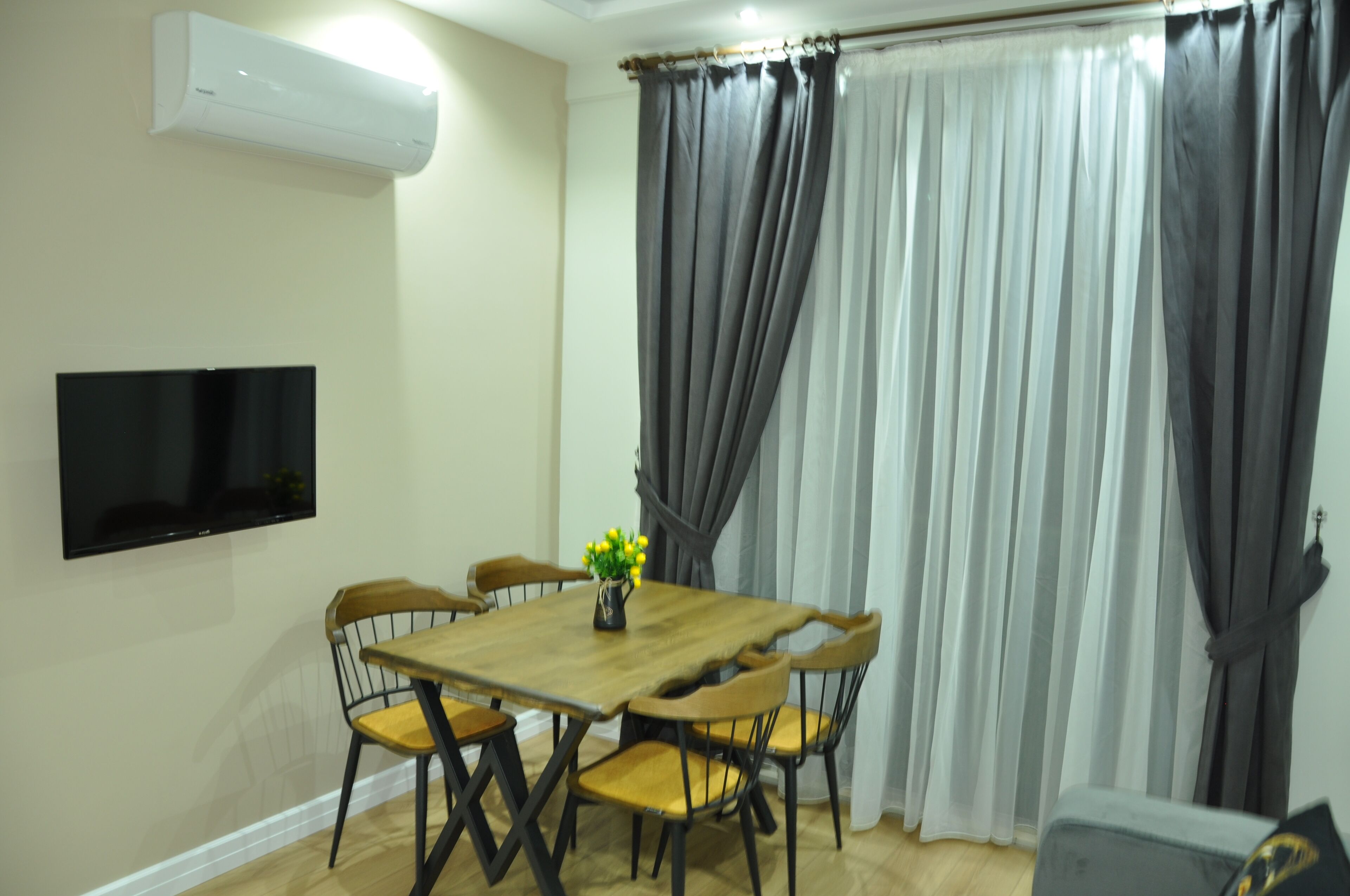 A dining area with a wooden table, six chairs, a flat-screen TV mounted on the wall, and a window with grey curtains.